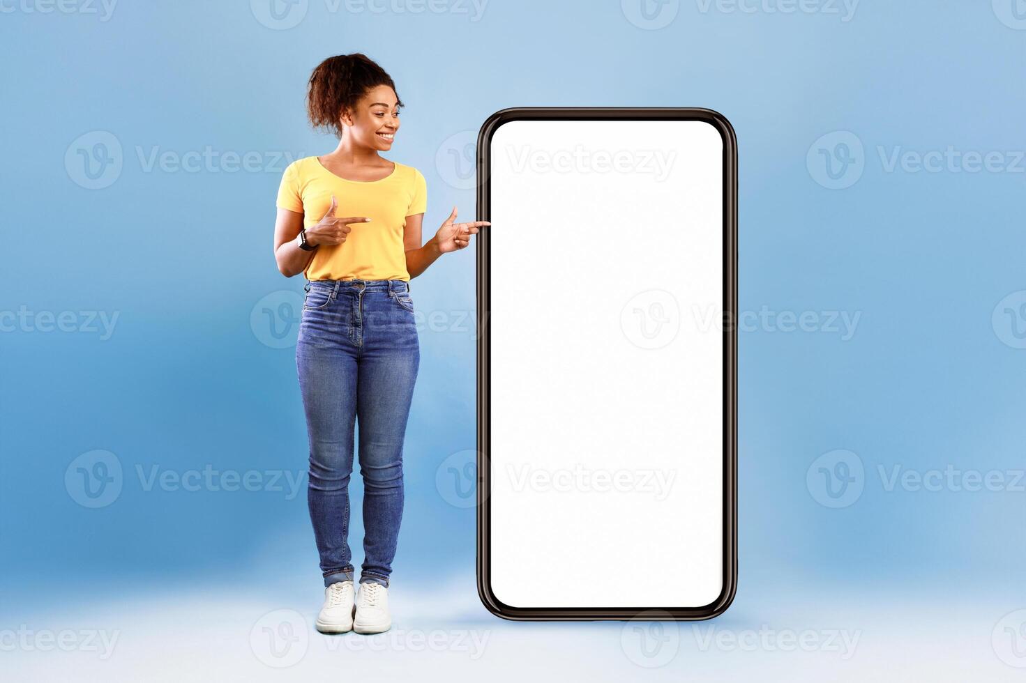 Cheerful African American woman standing next to huge smartphone, pointing at empty screen, advertising new mobile application or website. Blue studio background, cellphone display mockup photo