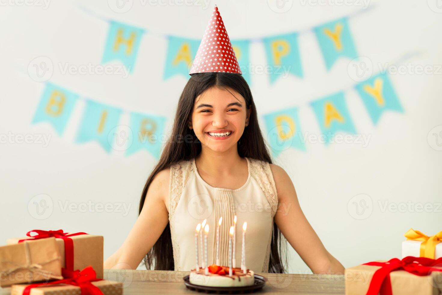 Portrait of cheery Indian teen girl in party hat sitting at table with gift boxes and birthday cake with candles at home. Joyful Asian adolescent celebrating holiday, looking at camera and smiling photo