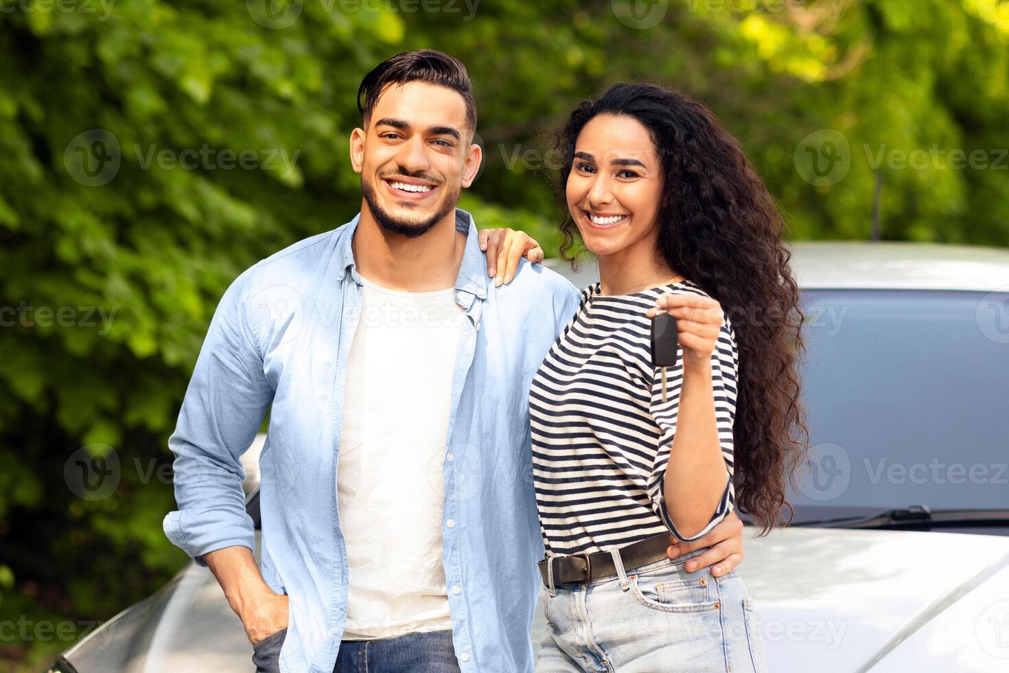 Cheerful beautiful middle-eastern couple young man and woman standing outside by nice auto, hugging and showing key from automobile, having car trip together. Car renting, leasing, sharing concept photo