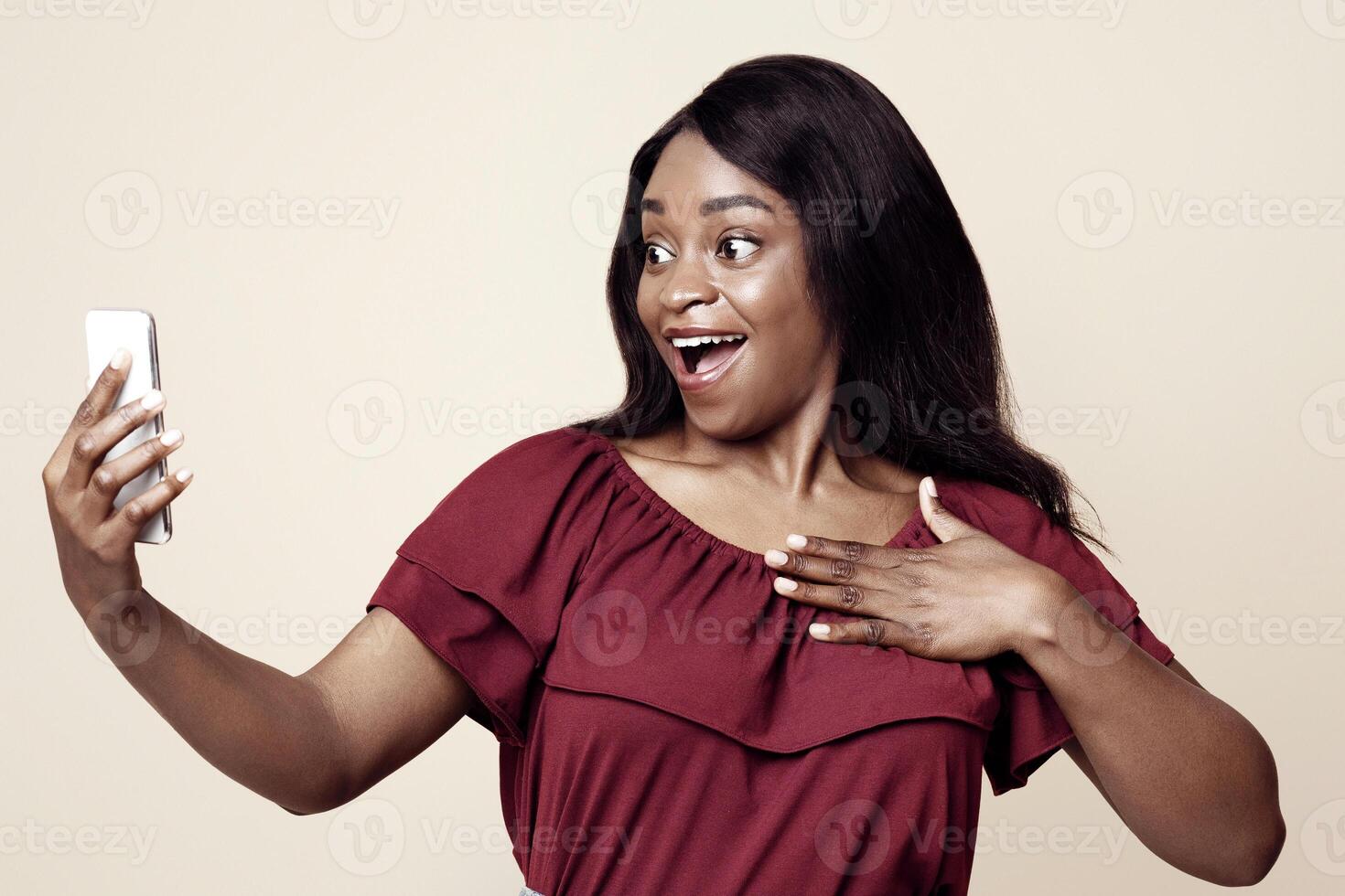 Surprised attractive millennial black woman having chat with friends, using mobile phone, showing excitement, happy to see her fellows, beige studio background, copy space photo