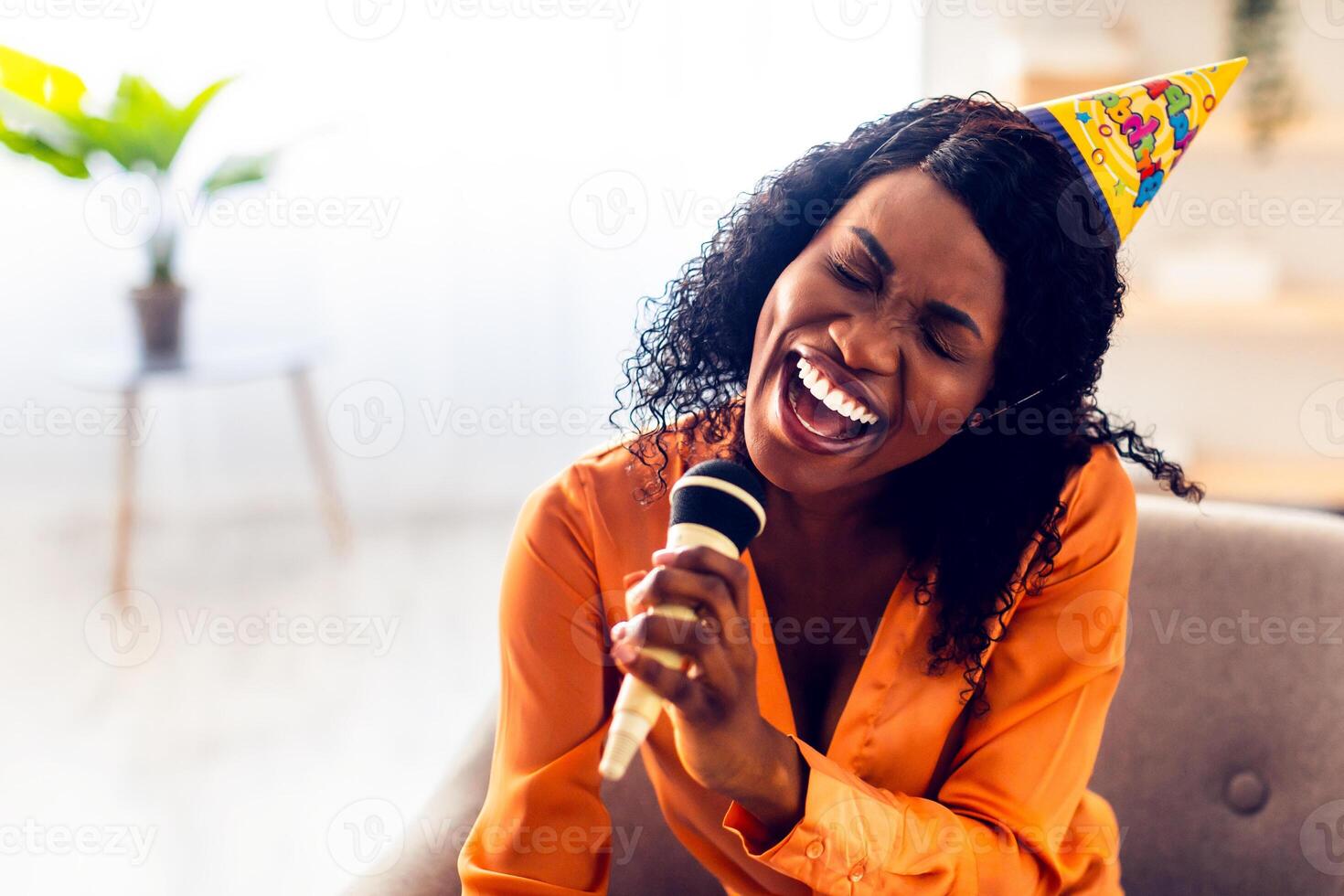Birthday Fun. African American Woman Singing Song With Eyes Closed Holding Microphone Having Karaoke Party Sitting In Chair At Home, Wearing Festive Hat. B-Day Leisure Concept photo