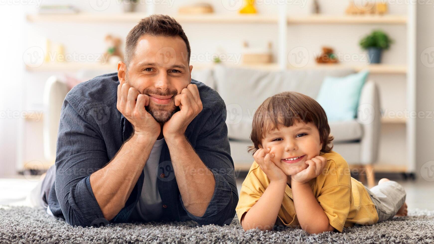 Happy caucasian family. Portrait of cute little boy and his daddy smiling to camera, lying together on floor at home, leaning heads on hands, empty space photo