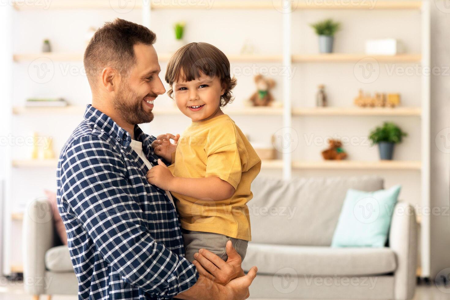 Happy childhood. Cute little baby boy smiling to camera, enjoying connection with daddy, happy man looking at son with love, carrying him on hands at home photo