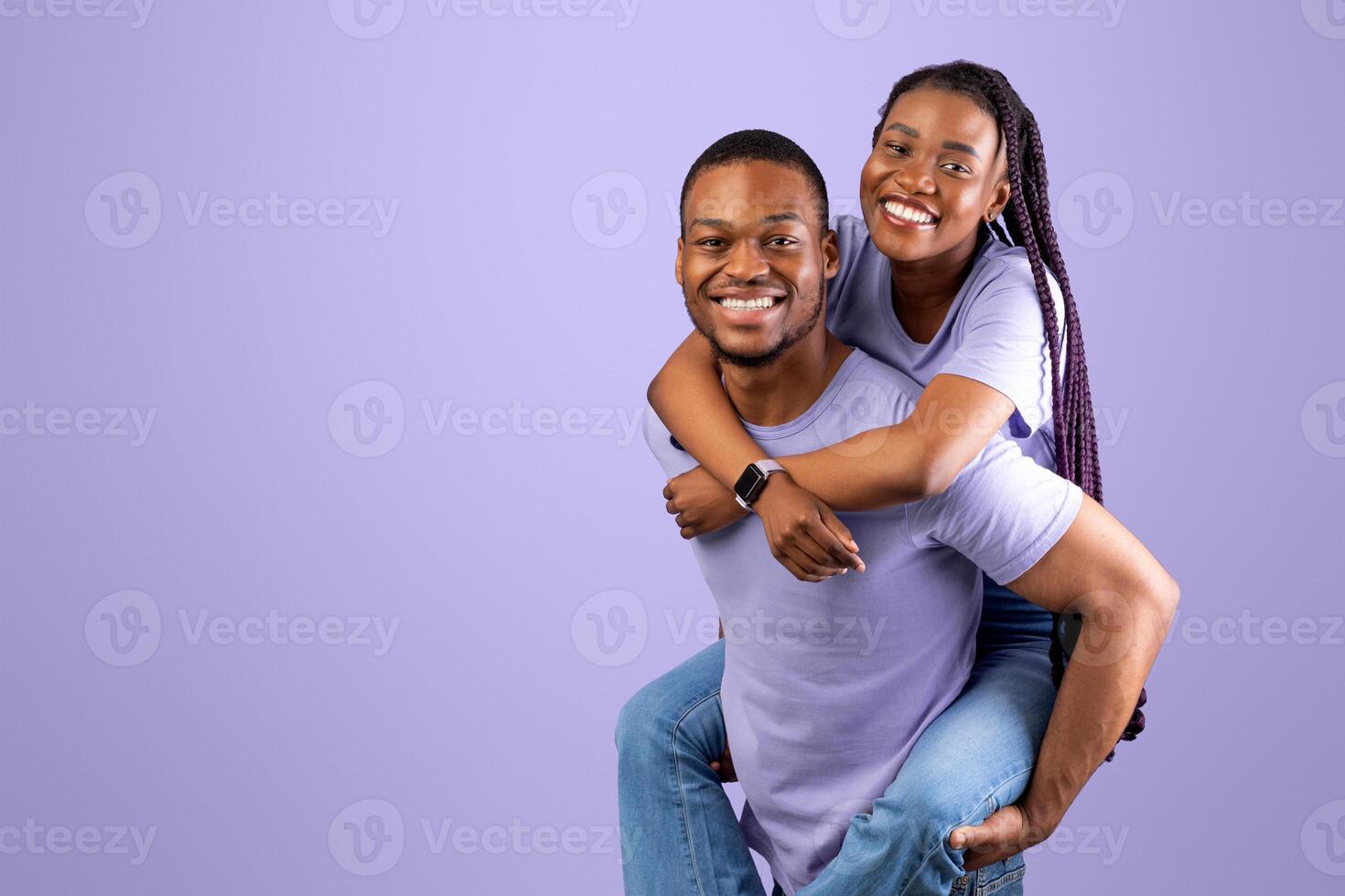 Piggyback Ride. Portrait of smiling African American man carrying and riding his woman on back, enjoying time together, standing isolated over purple studio background, having fun fooling around photo