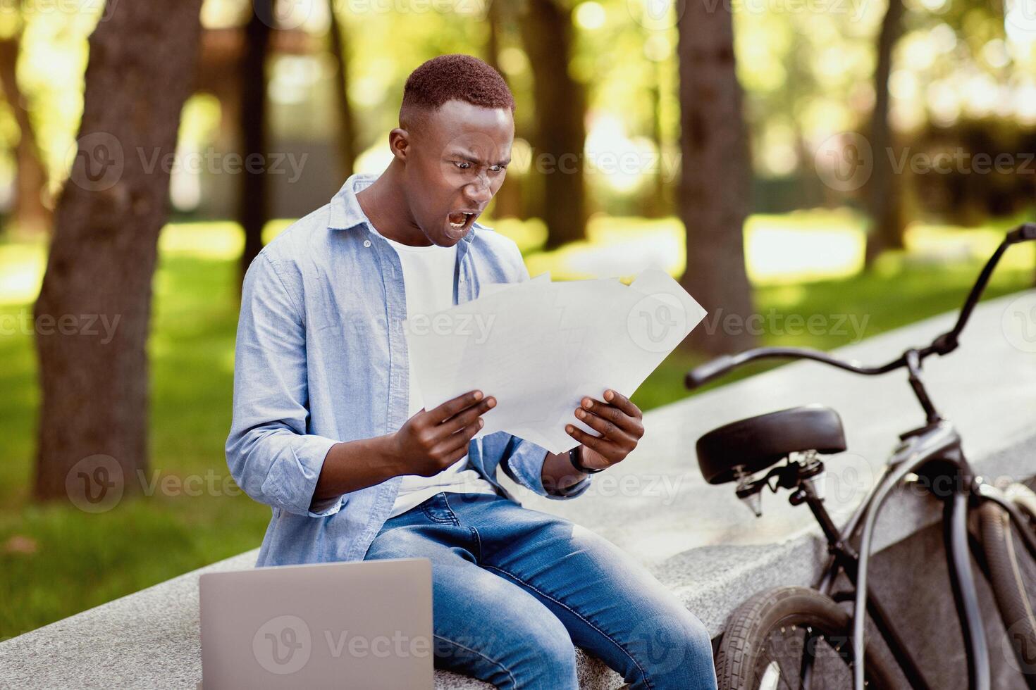 Online job concept. Young black freelancer with bike and laptop finding mistake in his work project, shouting in terror at park. Millennial African American guy having problem with documents outside photo