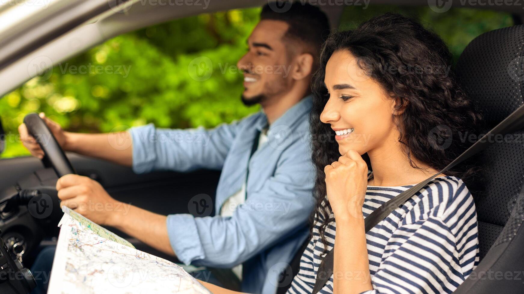 Happy middle-eastern lovers going vacation by car, handsome guy driving nice auto, pretty long-haired young woman looking at road map and smiling, side view. Navigation, travelling, trip concept photo