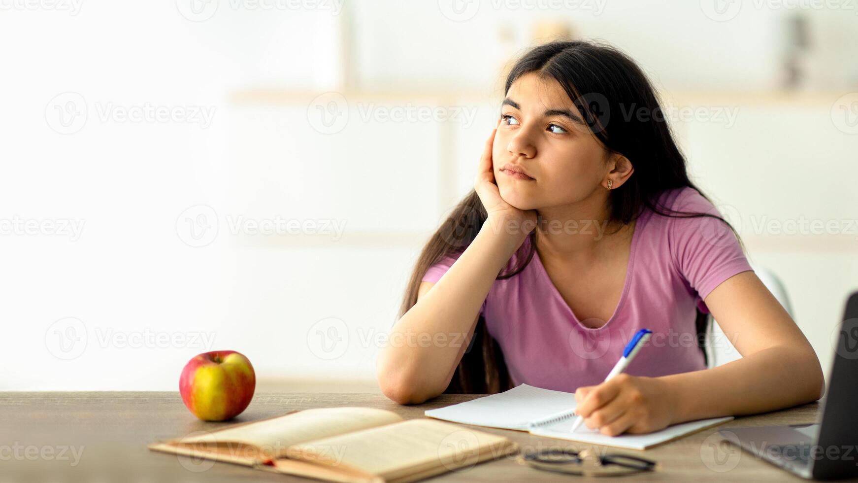 Thoughtful Indian teen girl studying online from home, taking notes during web lesson, feeling bored at home. Pensive adolescent participating in dull lecture, learning remotely during covid lockdown photo