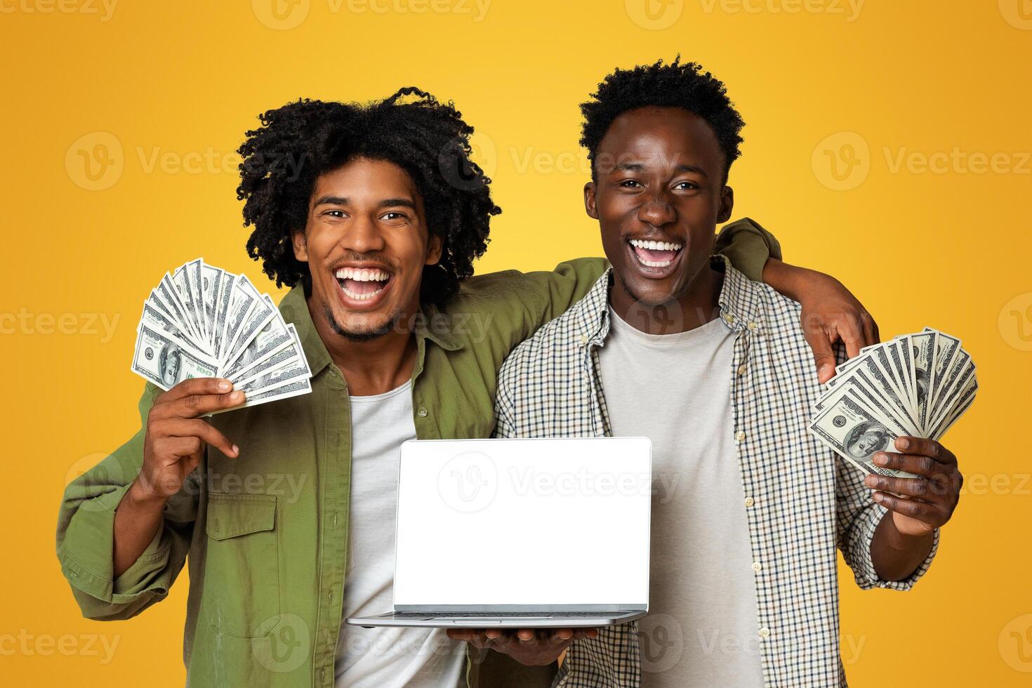 Online Lottery. Two black guys holding dollar cash and laptop with blank white screen, happy african american friends with recommending new internet website while standing on beige background, mockup photo