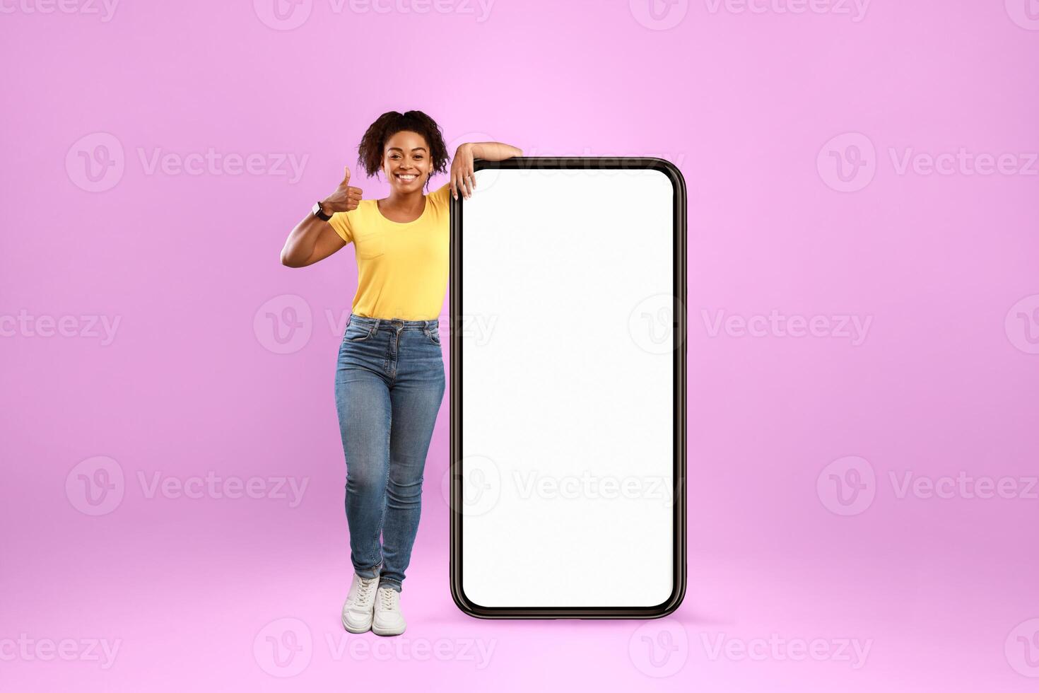 Advertisement for mobile application. Cheery African American lady standing near modern smartphone with empty screen and showing thumb up gesture on violet studio background, mockup photo