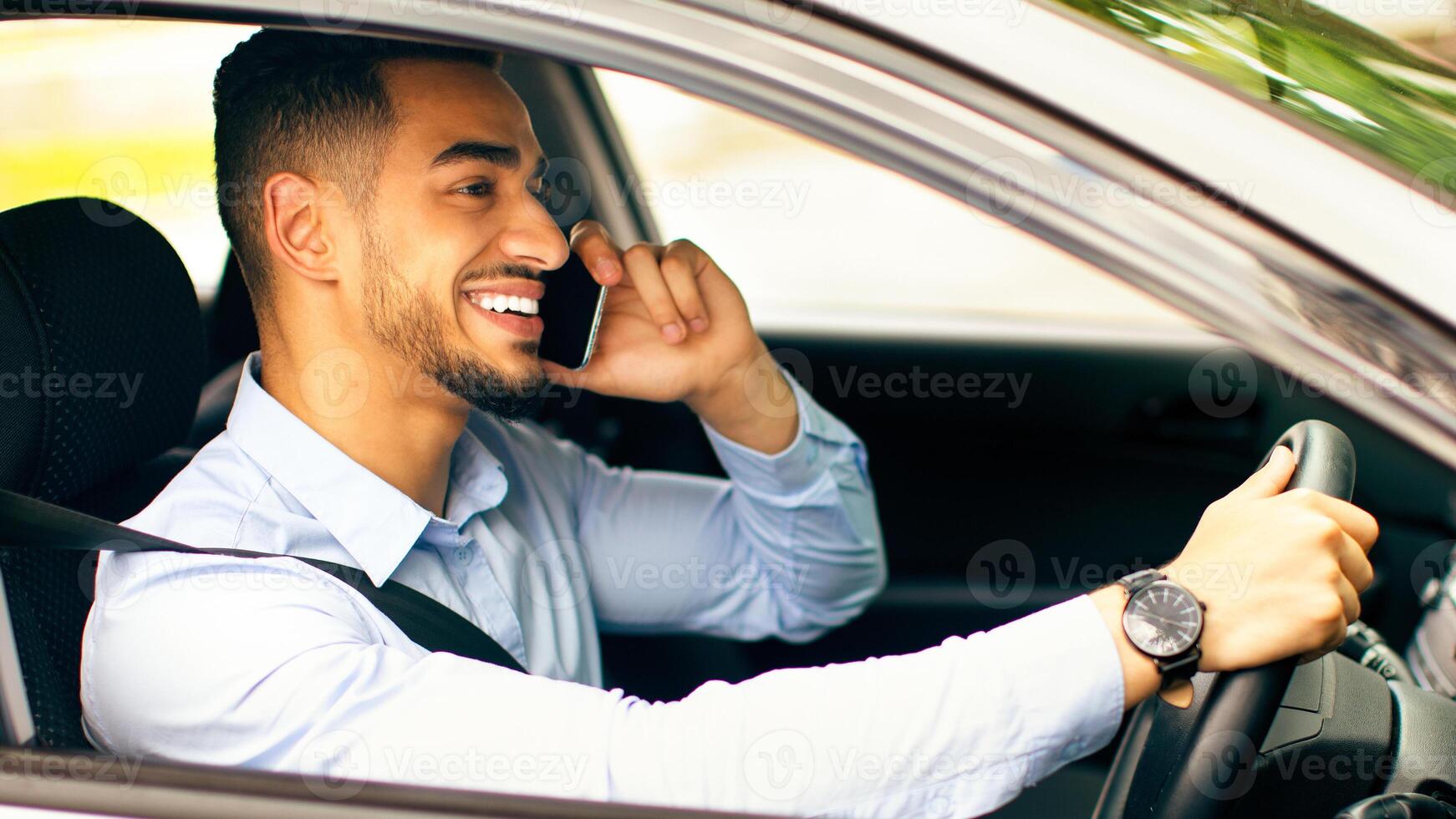 Bad driving habits, unsafe driving, driving and gadgets concept. Cheerful arab man entrepreneur going to office by auto, talking on phone while driving car, side view, copy space photo