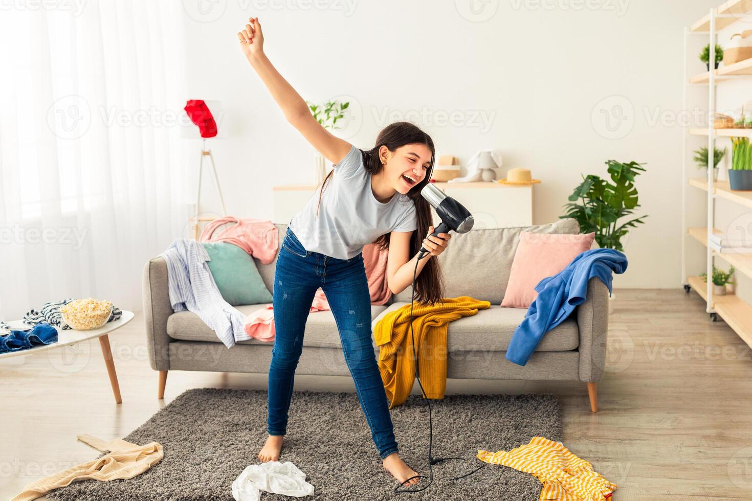 Full length of Indian teenage girl dancing and singing in messy room, using blowdryer as mic, having home concert. Adorable adolescent having fun, entertaining herself during coronavirus quarantine photo