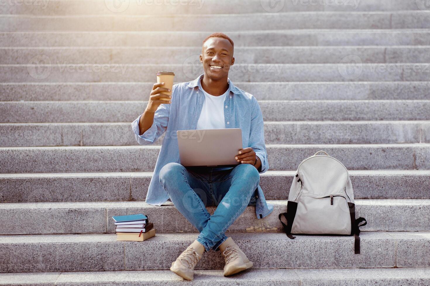 Online communication. Young African American man with takeout coffee talking to someone via internet on stairs outside photo