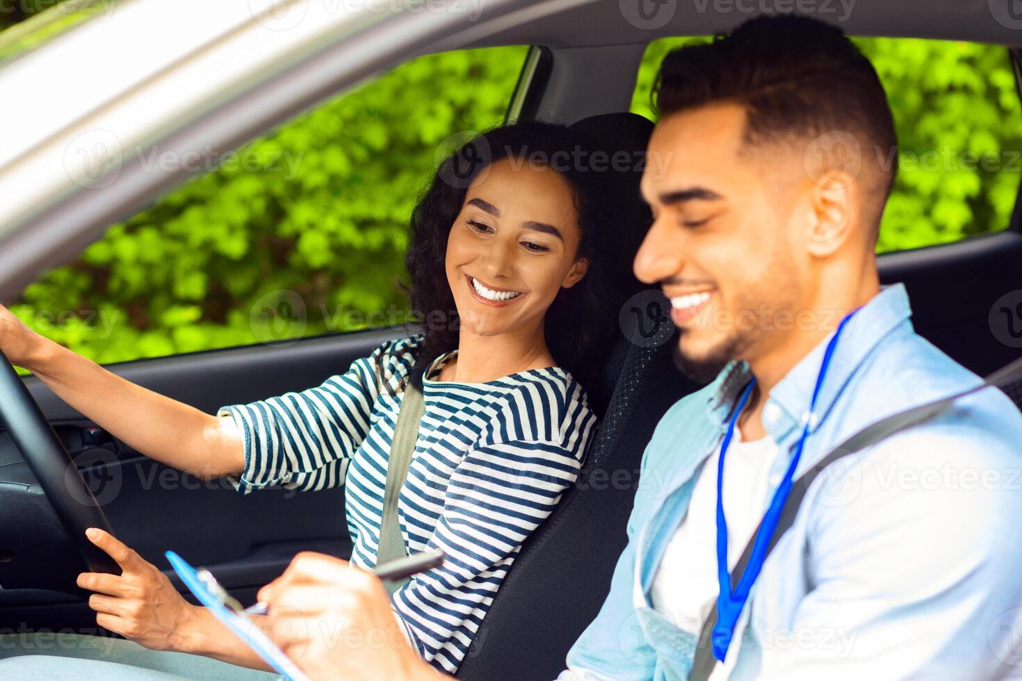 Cheerful middle-eastern guy instructor examinating happy brunette lady student, taking notes at chart while sitting by young woman driving car and waiting for test results, side view, copy space photo