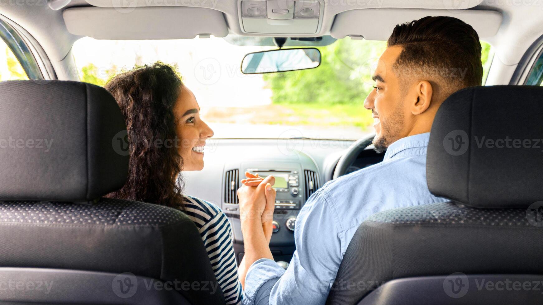 Loving multiracial couple holding hands and smiling to each other while sitting in comfortable auto, back view. happy arab guy having car trip with his beautiful girlfriend, summer vacation concept photo