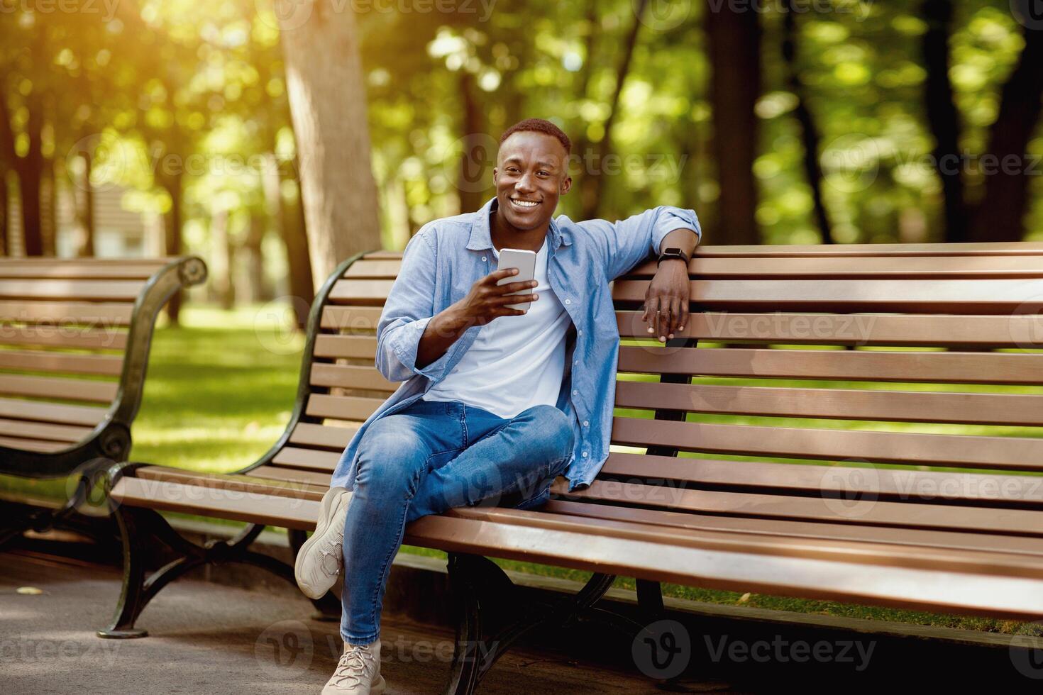 Smiling black man with mobile phone relaxing on bench at park on beautiful summer day photo