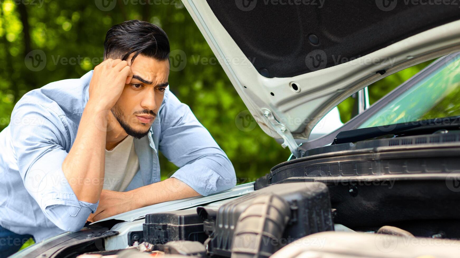 Puzzled arab man leaning on open car hood, looking at engine, having automobile crush in the middle of the road. Nervous middle-eastern guy driver waiting for car service, closeup photo