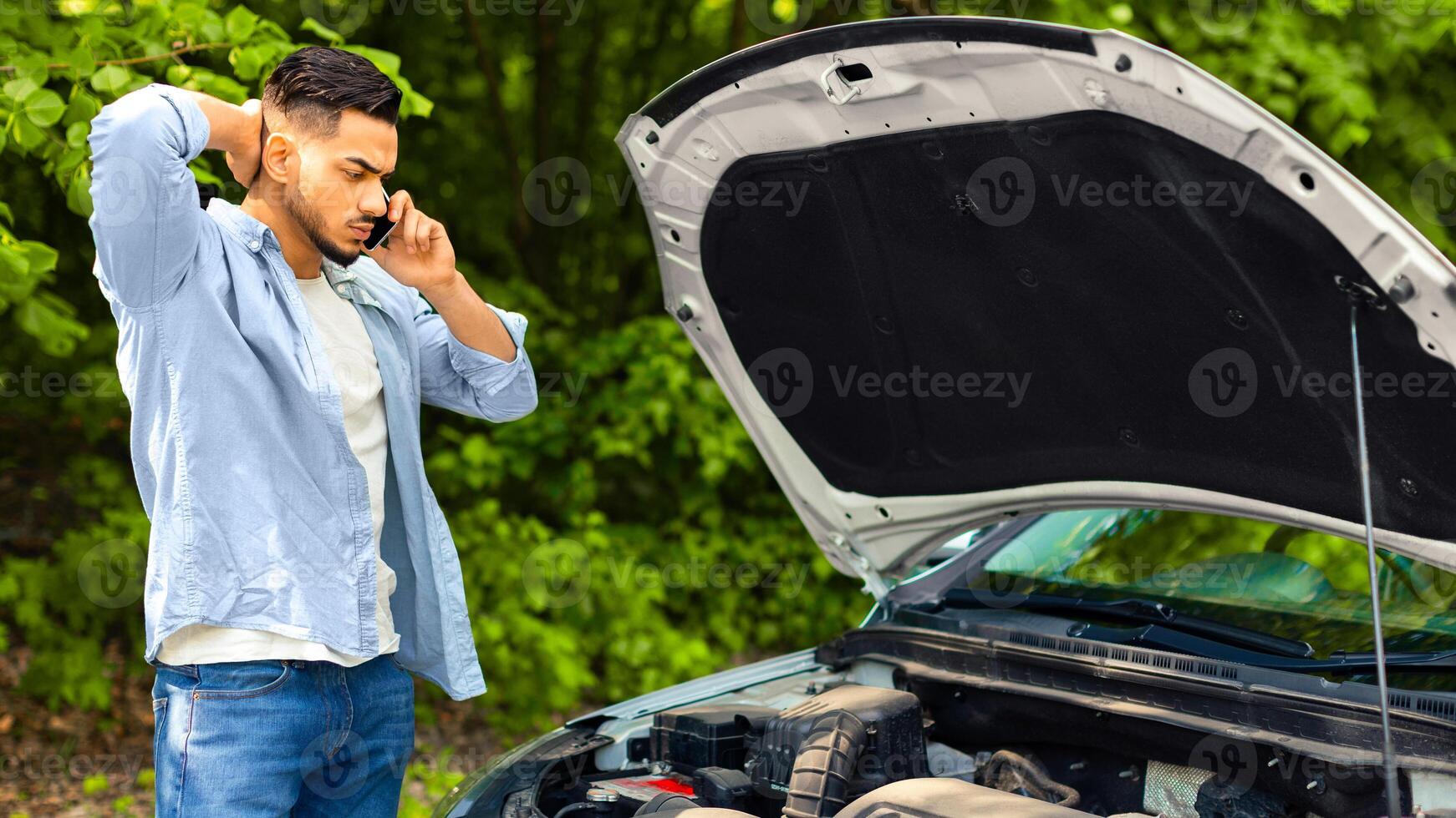 Sad arab man looking at car engine and calling to auto service, got broken at highway on a way to countryside. Middle-eastern guy having phone conversation with friends, asking for help with auto photo