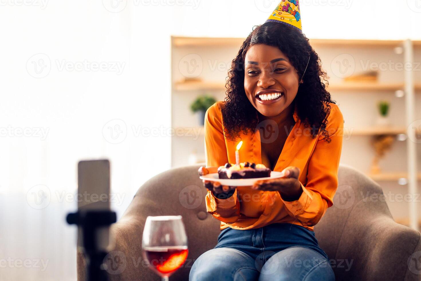 Happy African Female Holding Birthday Cake Making Wish Near Phone Having Call, Celebating Holiday Online In Living Room Indoor. Distance B-Day Celebration Concept. Selective Focus photo