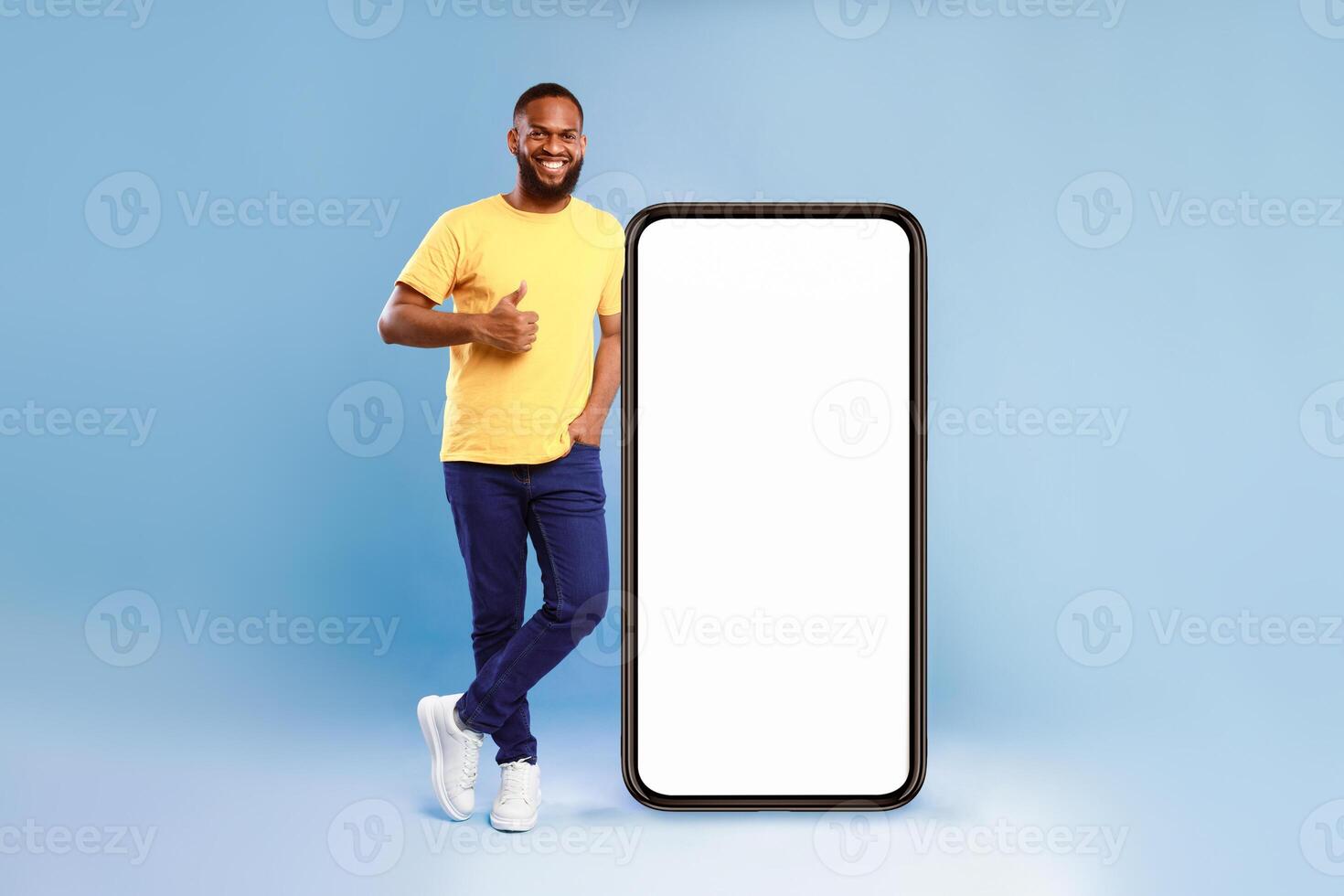 Cool mobile application. Joyful African American guy in gesturing thumb up near giant smartphone over blue studio background, mockup for website or new app on blank screen photo