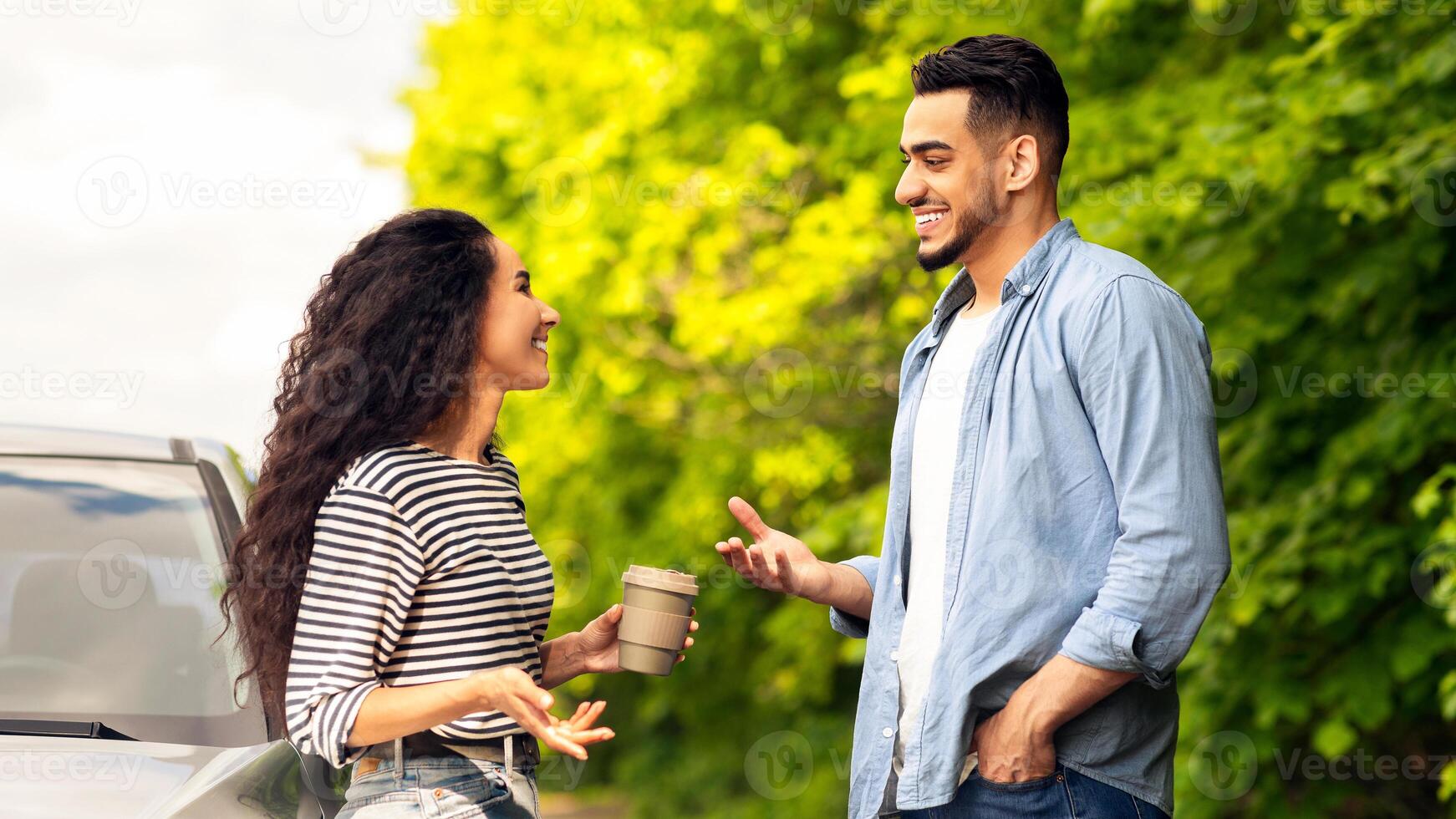 Joyful lovers having break while travelling by auto together, beautiful young middle-eastern man and woman standing by car at country side, drinking coffee and having conversation, side view photo