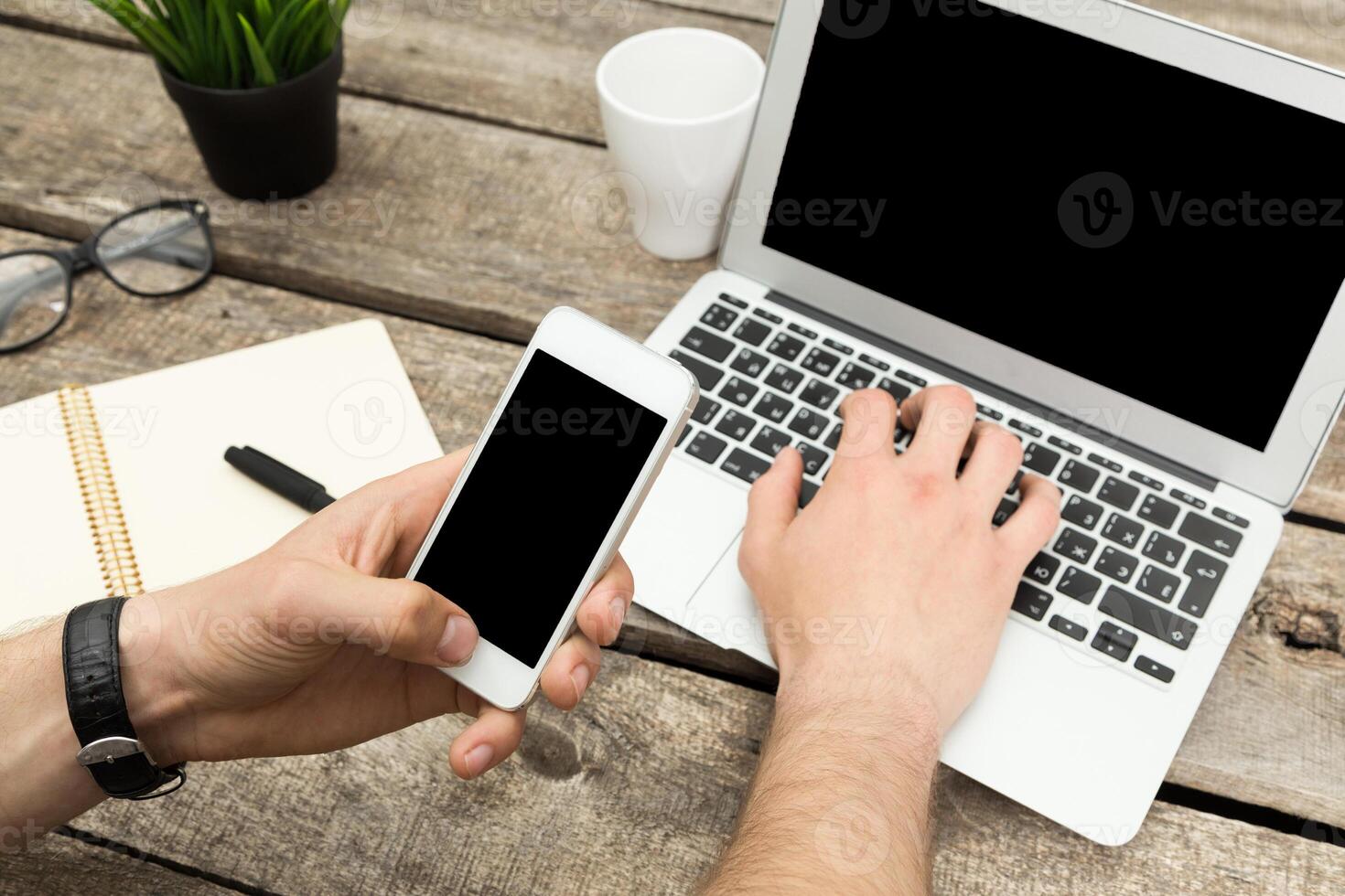 Hands using smartphone and laptop on wooden table in a casual workspace setting photo