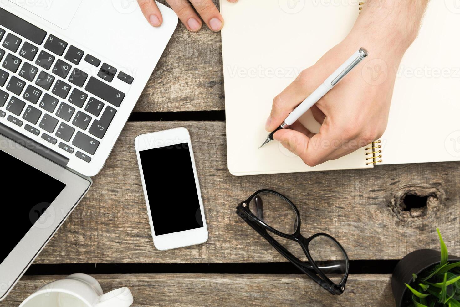 Person writing notes while using a laptop and mobile phone on a rustic wooden table during a work session photo