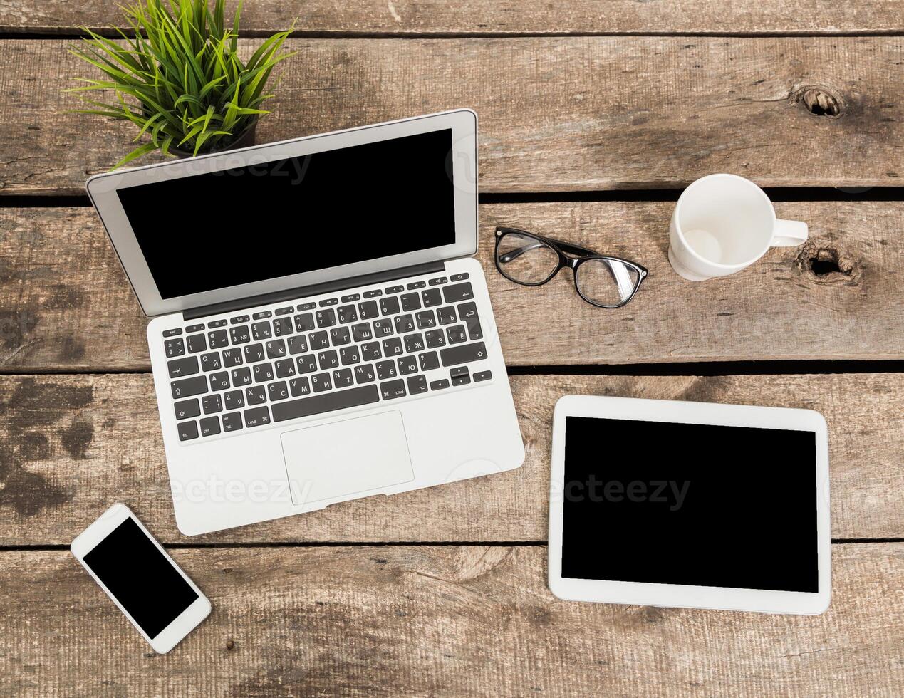 Workspace setup on wooden table with laptop, tablet, smartphone, glasses, and coffee cup during daytime photo