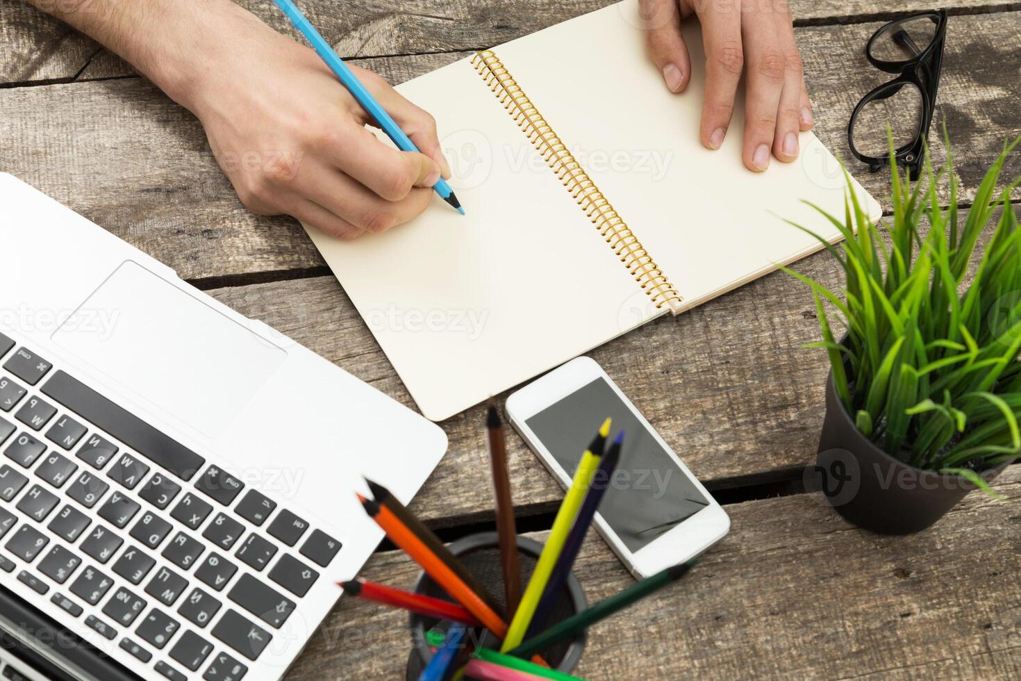 Writing in a notebook with a laptop and colored pencils on a rustic wooden table during a productive workspace session photo