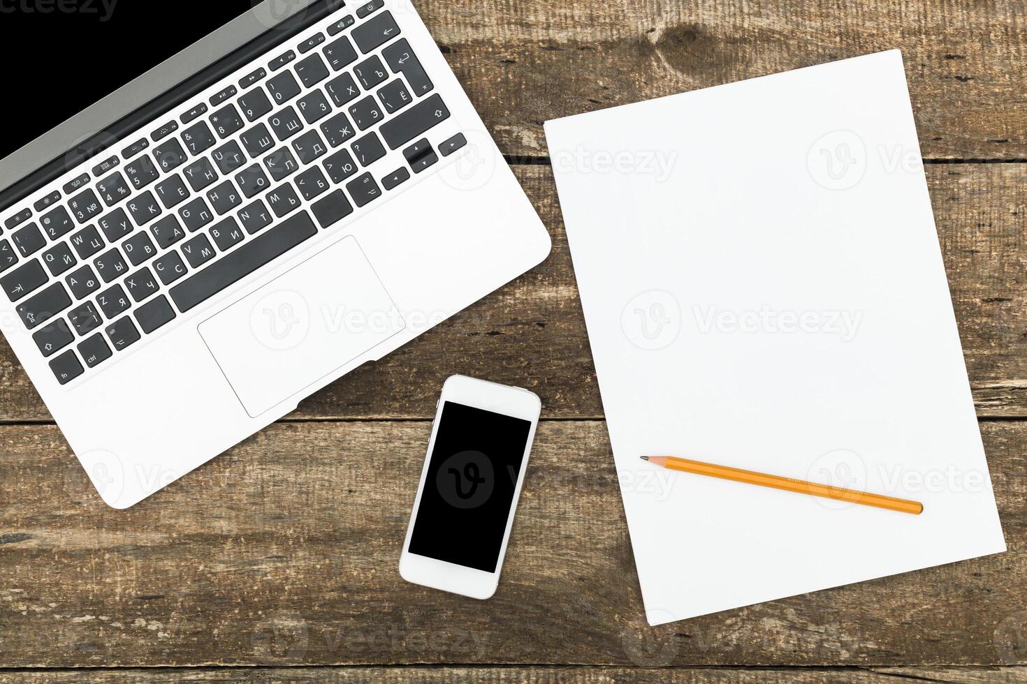 Workspace setup featuring a laptop, smartphone, blank paper, and pencil on a rustic wooden table in a cozy environment photo