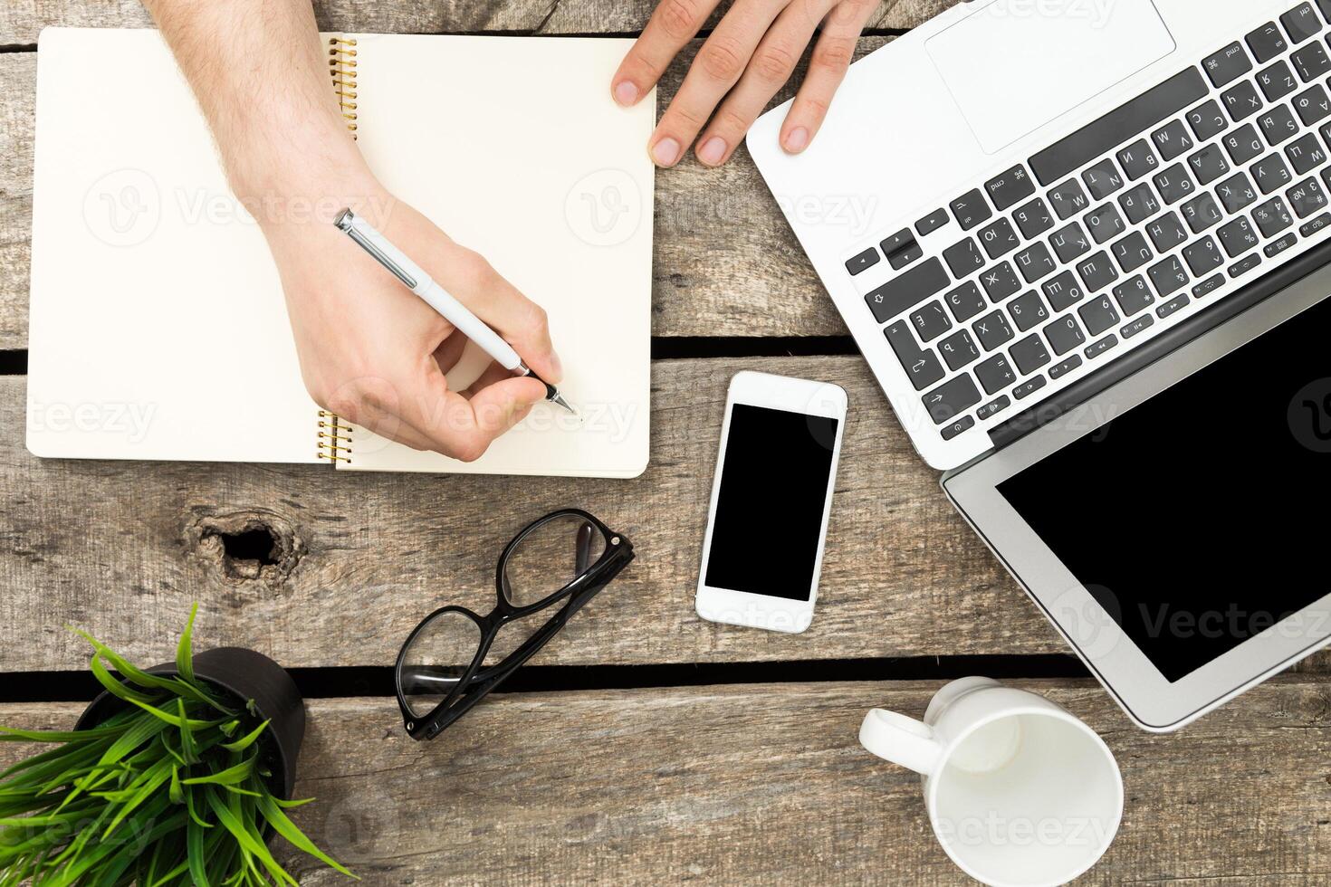 Writing notes while working on a laptop with a smartphone and coffee on a wooden table during daylight hours photo