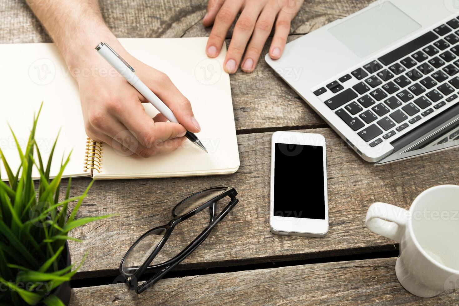 Person works on laptop while taking notes and planning at a wooden desk with a phone and coffee photo