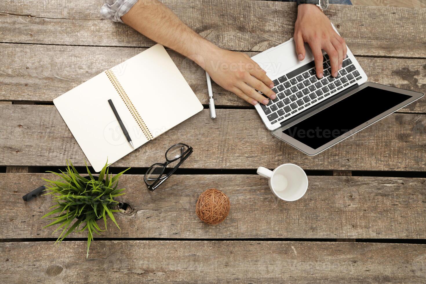 Person working on laptop at wooden table with notebook, glasses, and coffee cup during daylight hours photo