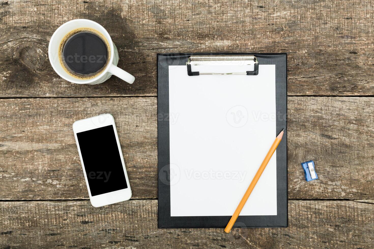Workspace with clipboard, coffee, smartphone, and pencil on rustic wooden table during morning hours photo