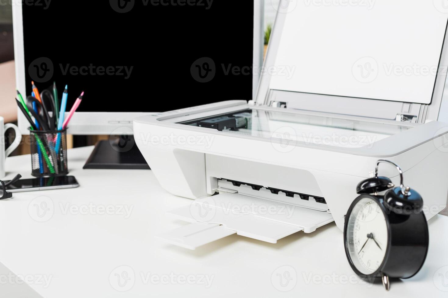 Office workspace featuring a printer, clock, and stationery arranged on a desk with a blank monitor in the background photo