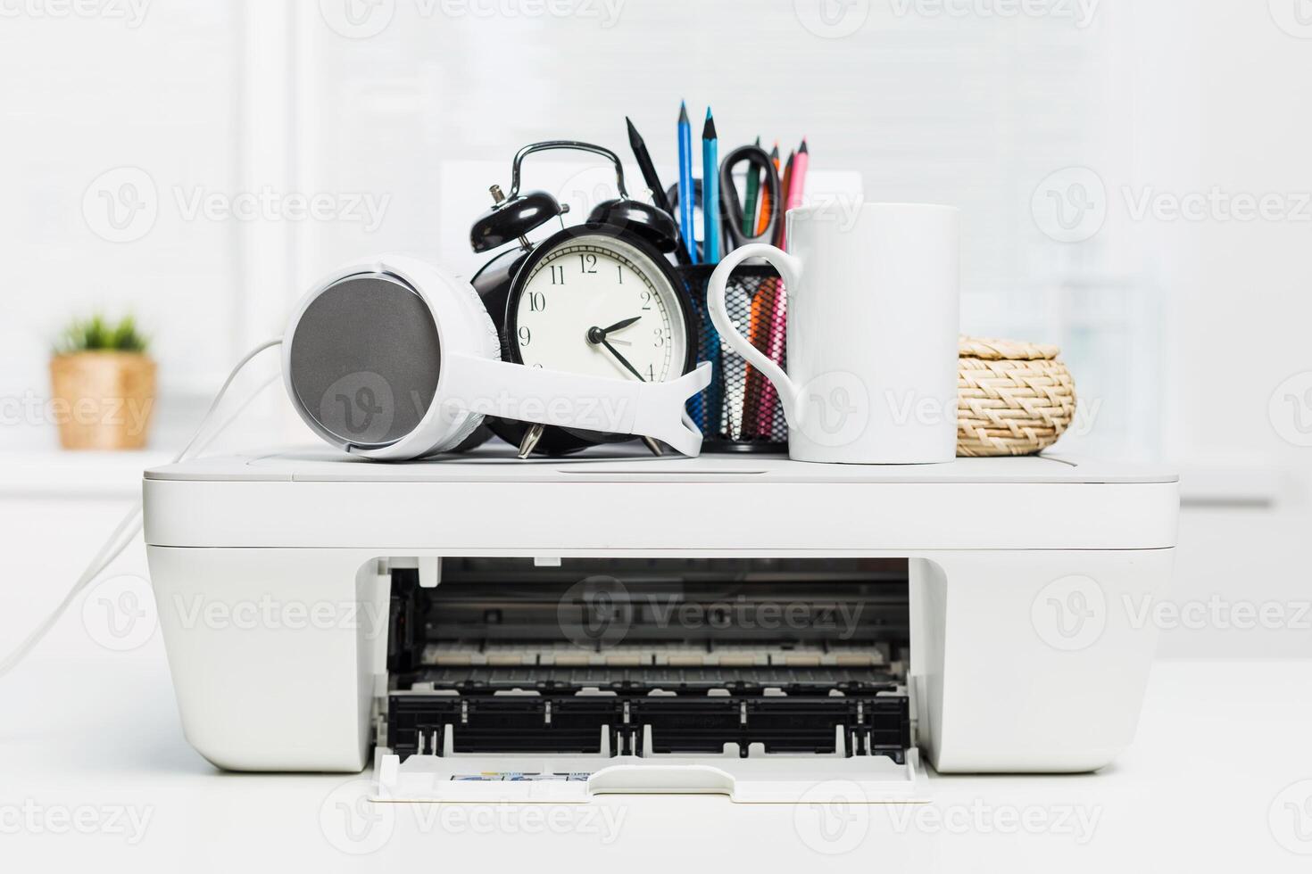 Office workspace with printer surrounded by stationery, clock, and cup during daylight hours photo