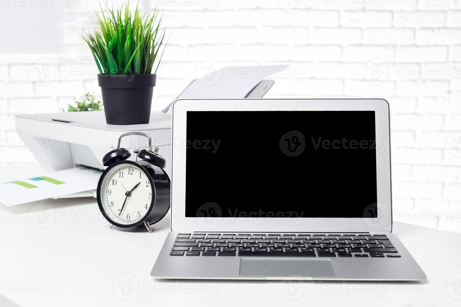 Modern workspace with a laptop, clock, printer, and greenery during daylight hours photo