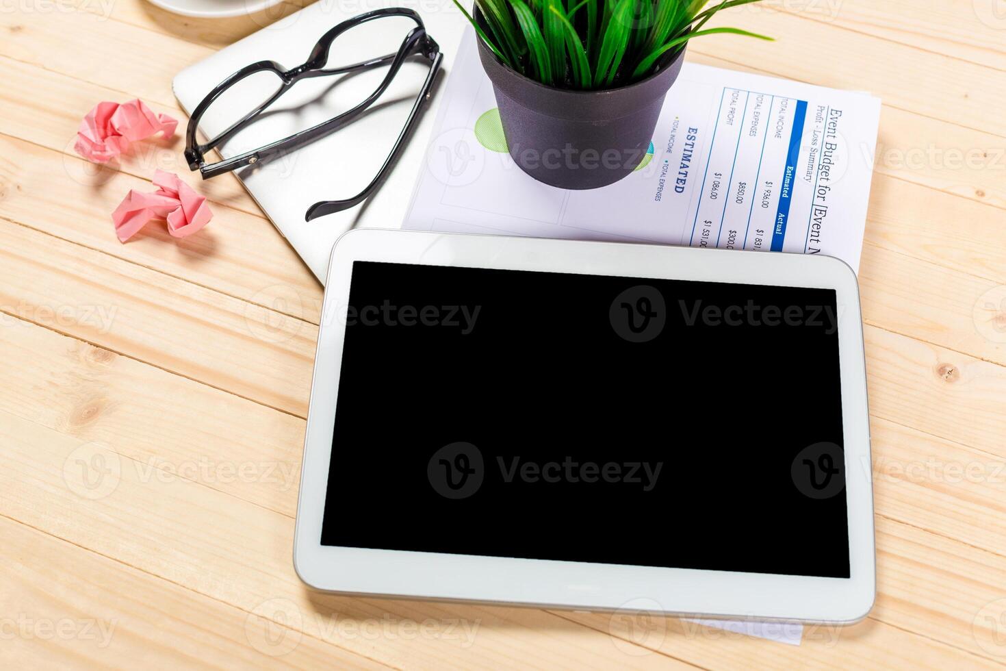 Workspace with tablet, documents, glasses, and plant on wooden table during daylight photo
