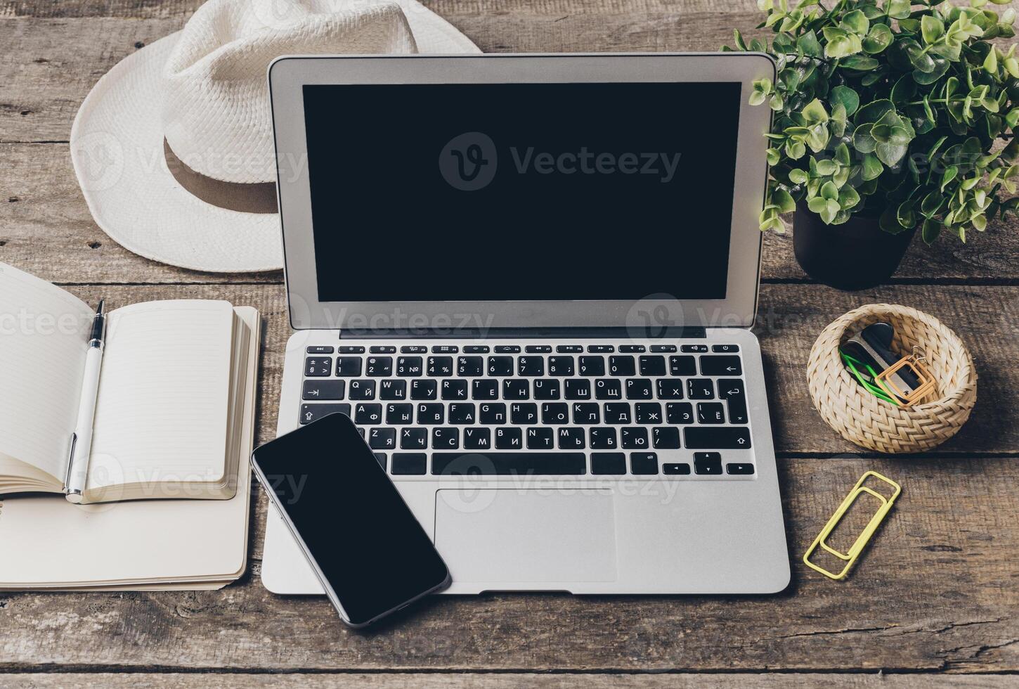 Workspace setup with a laptop, smartphone, notebook, and plant on a wooden table during daylight photo