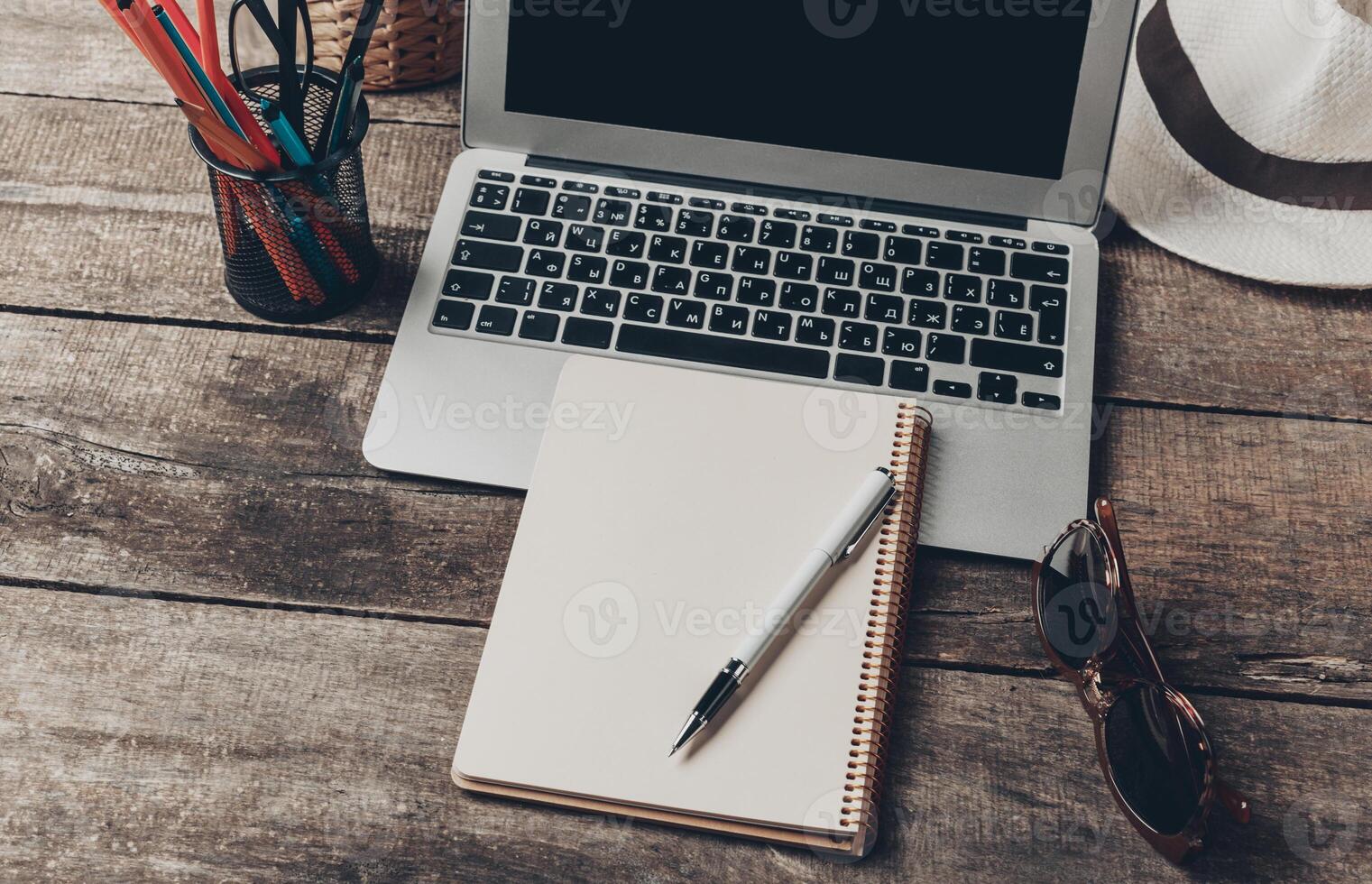 Work desk setup with laptop, notepad, pen, and sunglasses on a wooden table in a cozy environment photo