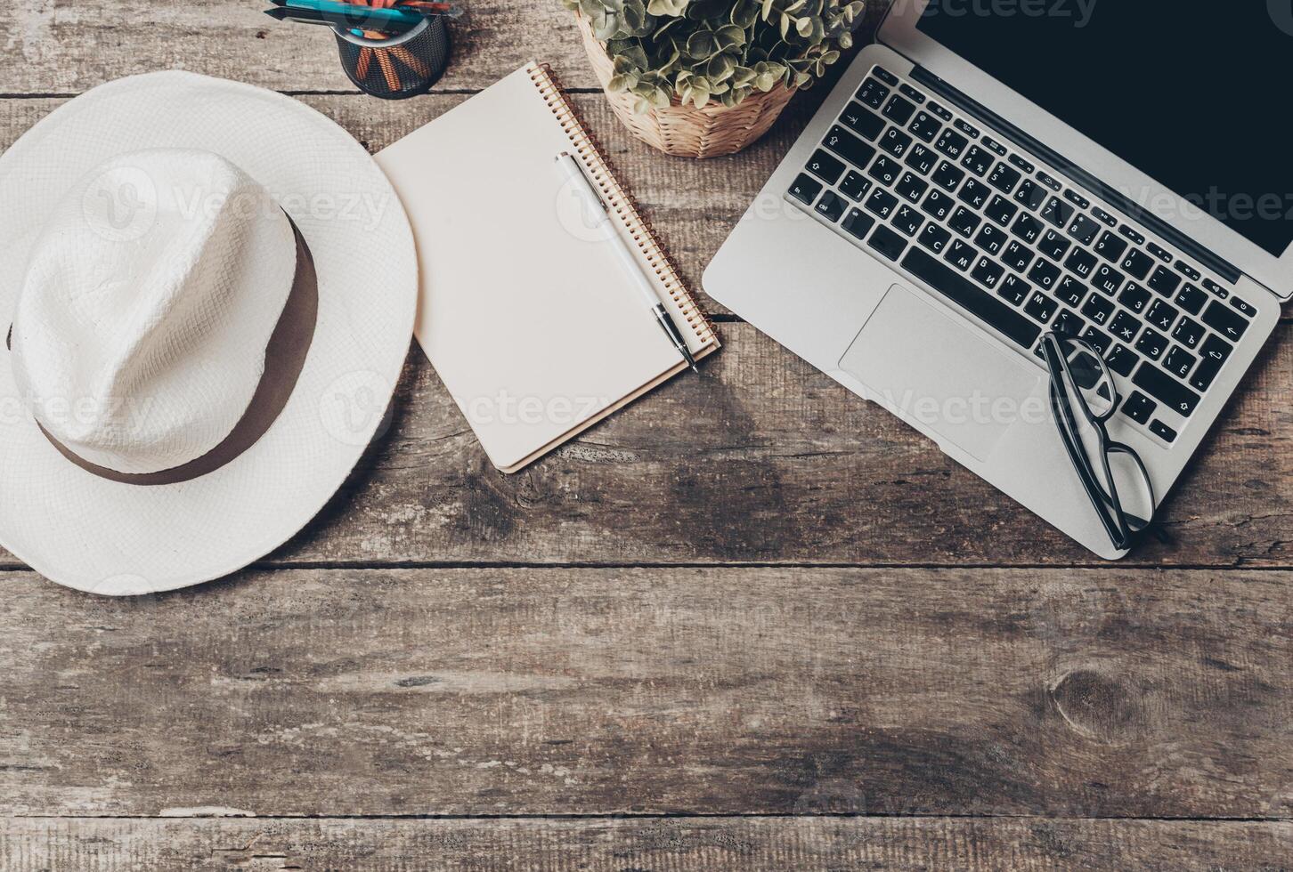 Workspace setup with a laptop, notebook, hat, and glasses on a rustic wooden table photo