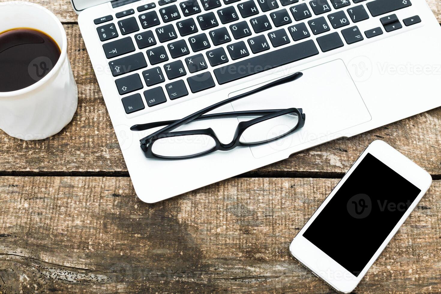 Workspace featuring a laptop, smartphone, glasses, and a cup of coffee set on a rustic wooden table during a productive afternoon photo
