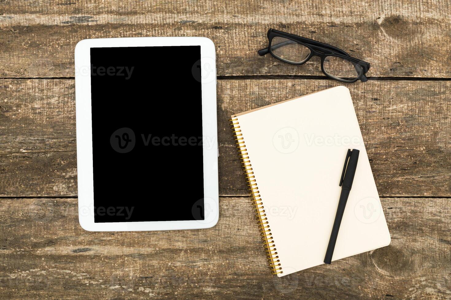 Workspace setup with tablet, notebook, pen, and glasses on rustic wooden table during daytime photo