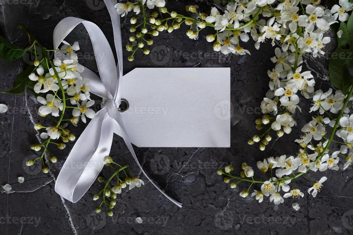A blank white card is tied with a bright yellow ribbon on the upper left corner. White blossoms frame the bottom and right edges of the card, set against a dark gray, textured surface, photo