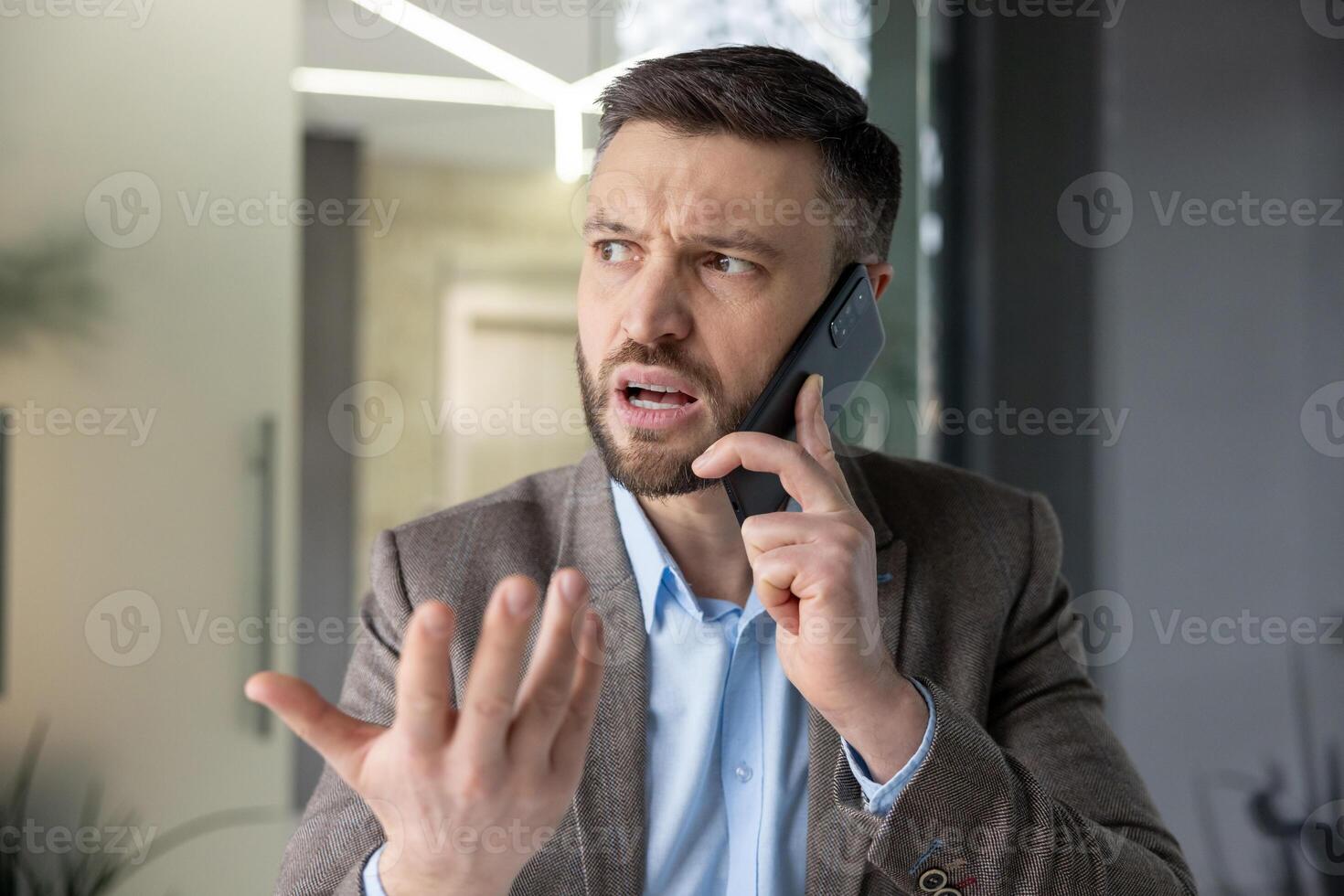 A frustrated businessman in a suit is engaged in a heated phone conversation, expressing his emotions with a hand gesture in an office setting. photo