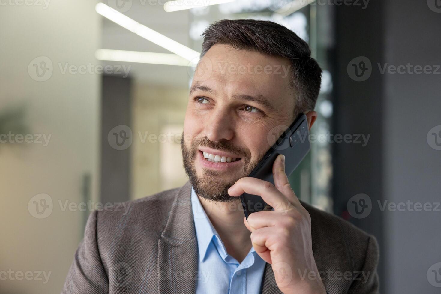 A confident businessman with a beard talking on his smartphone in a modern office setting, smiling while engaging in a conversation. photo