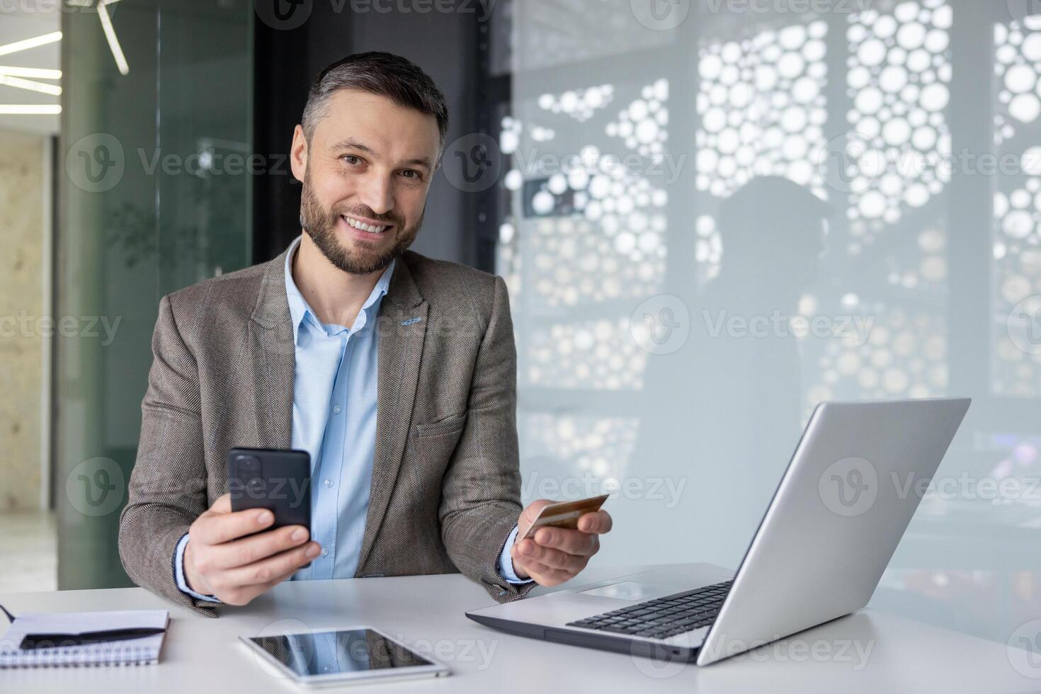 Smiling businessman sitting at desk with phone and credit card in modern office, showcasing online banking and digital payment. photo