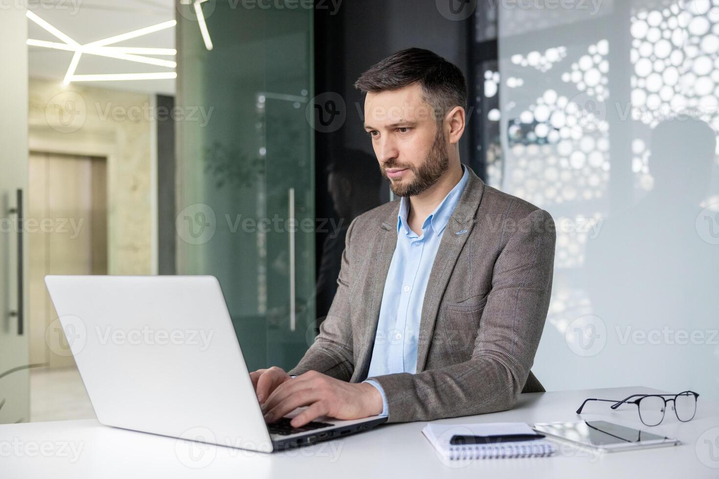 Professional businessman working on a laptop in a modern office setting, focused on his tasks. Background features glass walls and a notebook on the desk. photo