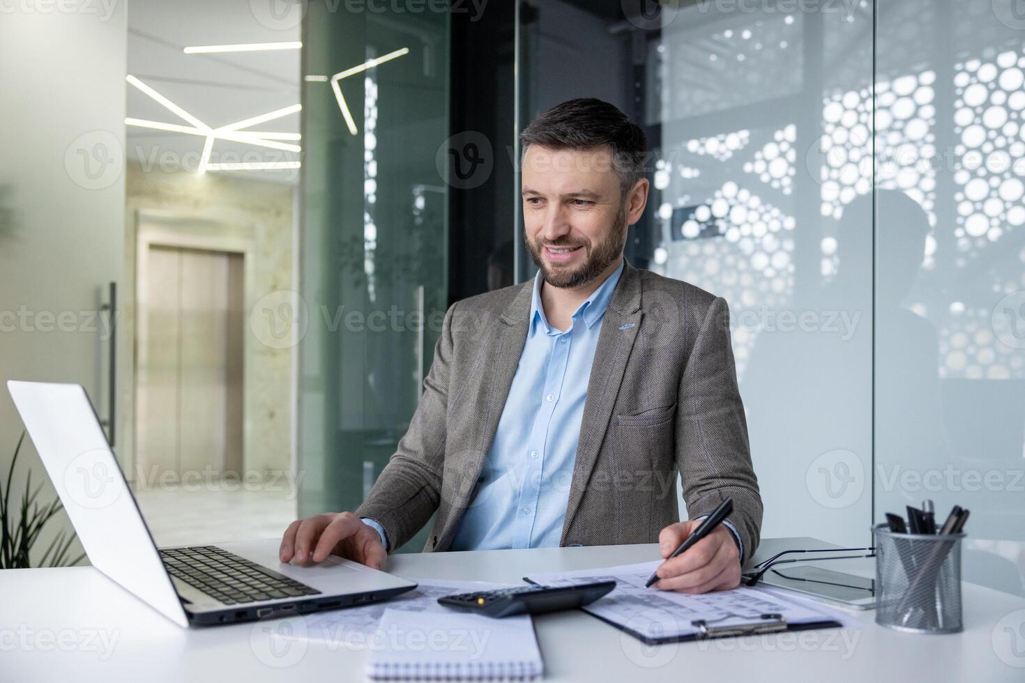 Confident businessman smiling while working on his laptop and taking notes in a sleek, modern office environment. photo