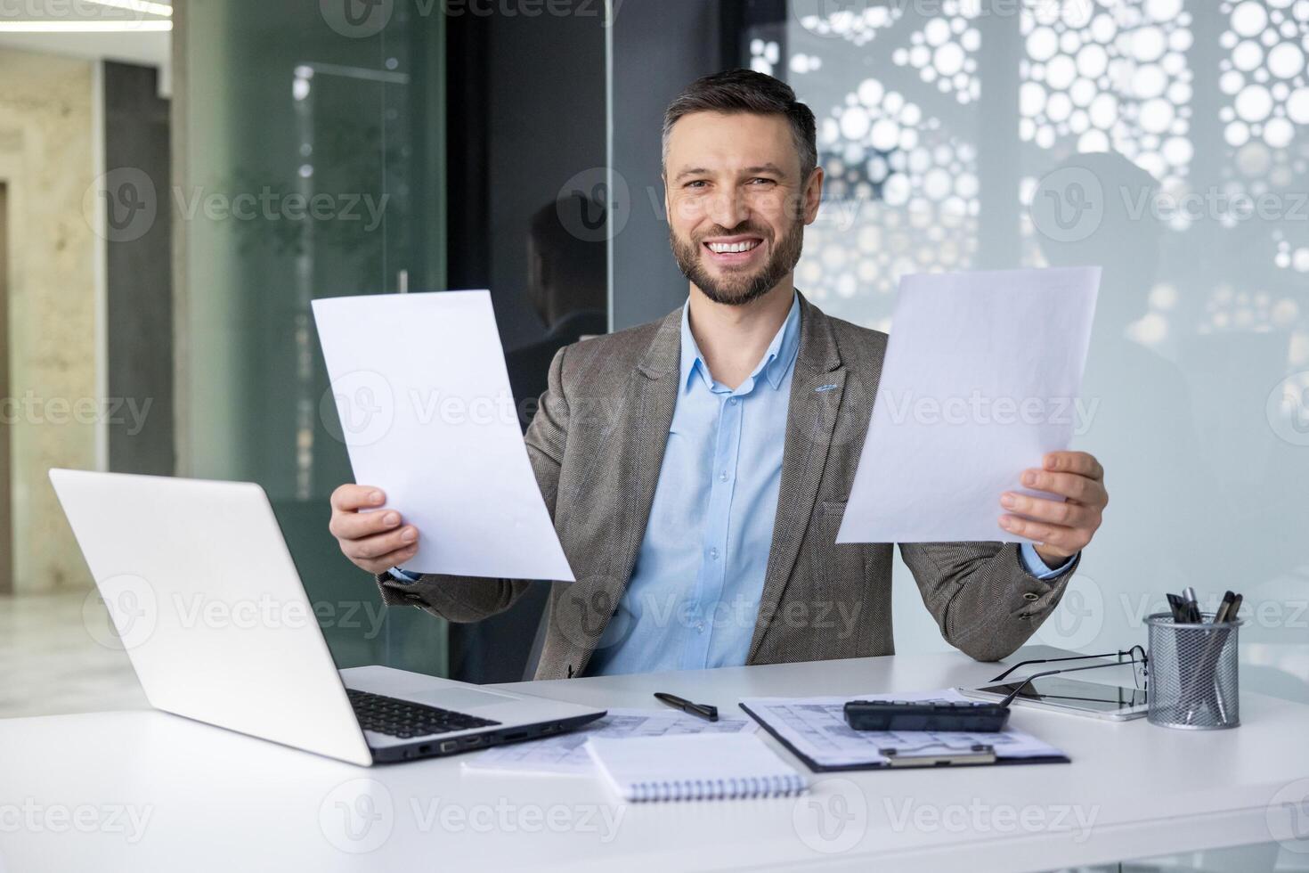 Confident smiling businessman showcasing documents at his desk in a modern office setting. Professional workspace with laptop and office supplies. photo