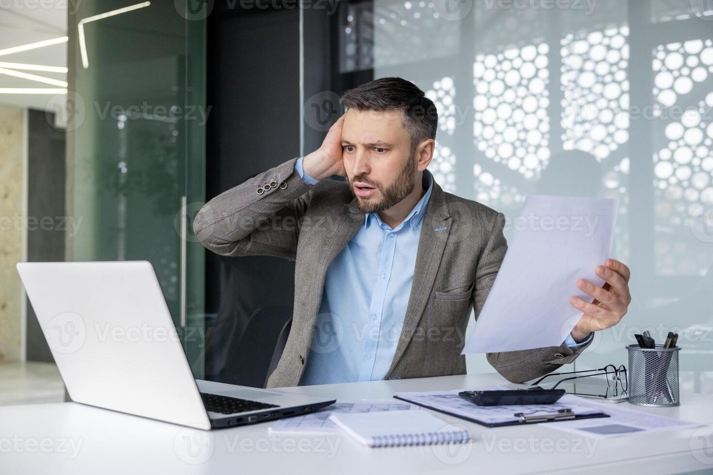Stressed businessman holding documents and working on a laptop in a modern office. Man is facing difficulty while analyzing data or dealing with financial problems. photo